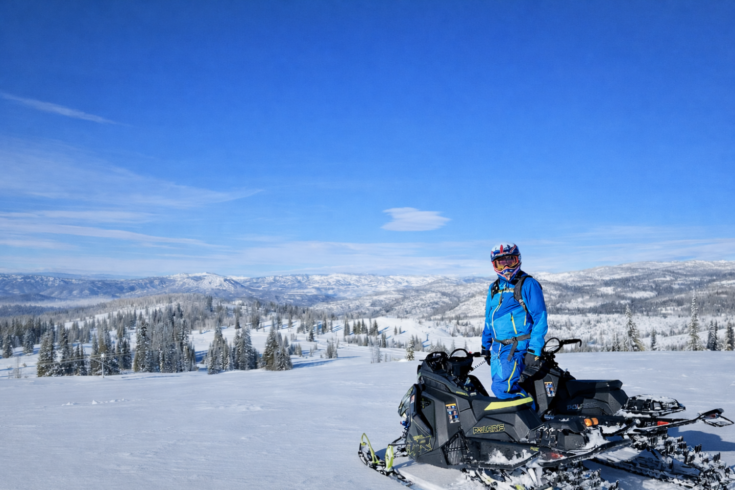 Snowmobiler in blue gear on a snowy mountain with clear blue sky and distant trees.