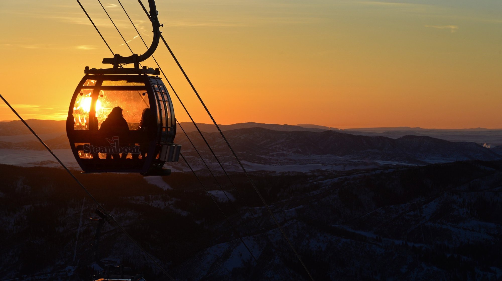 Gondola silhouetted against a sunset over snowy mountains.