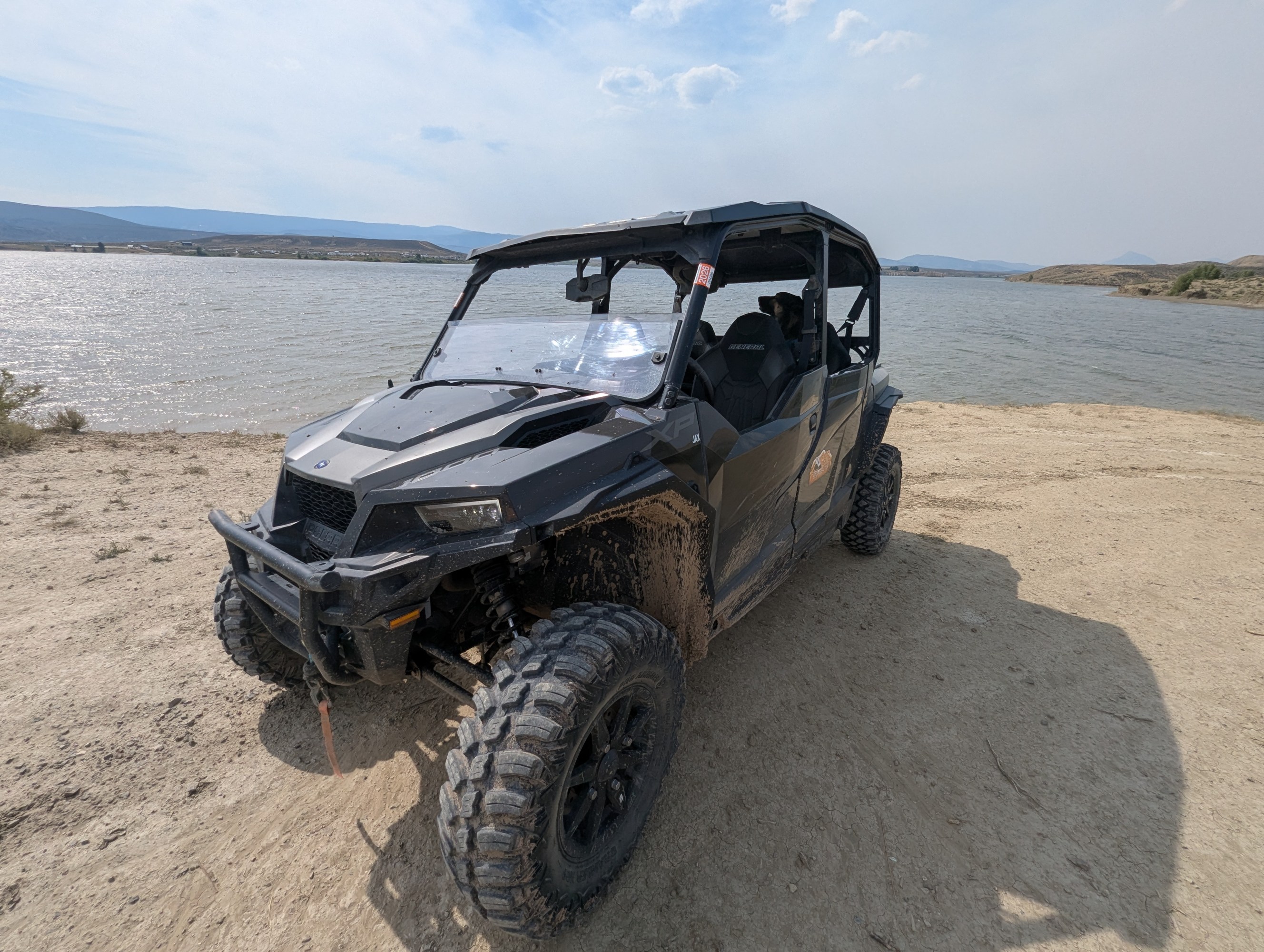 A muddy off-road vehicle parked by a lake on a sunny day.