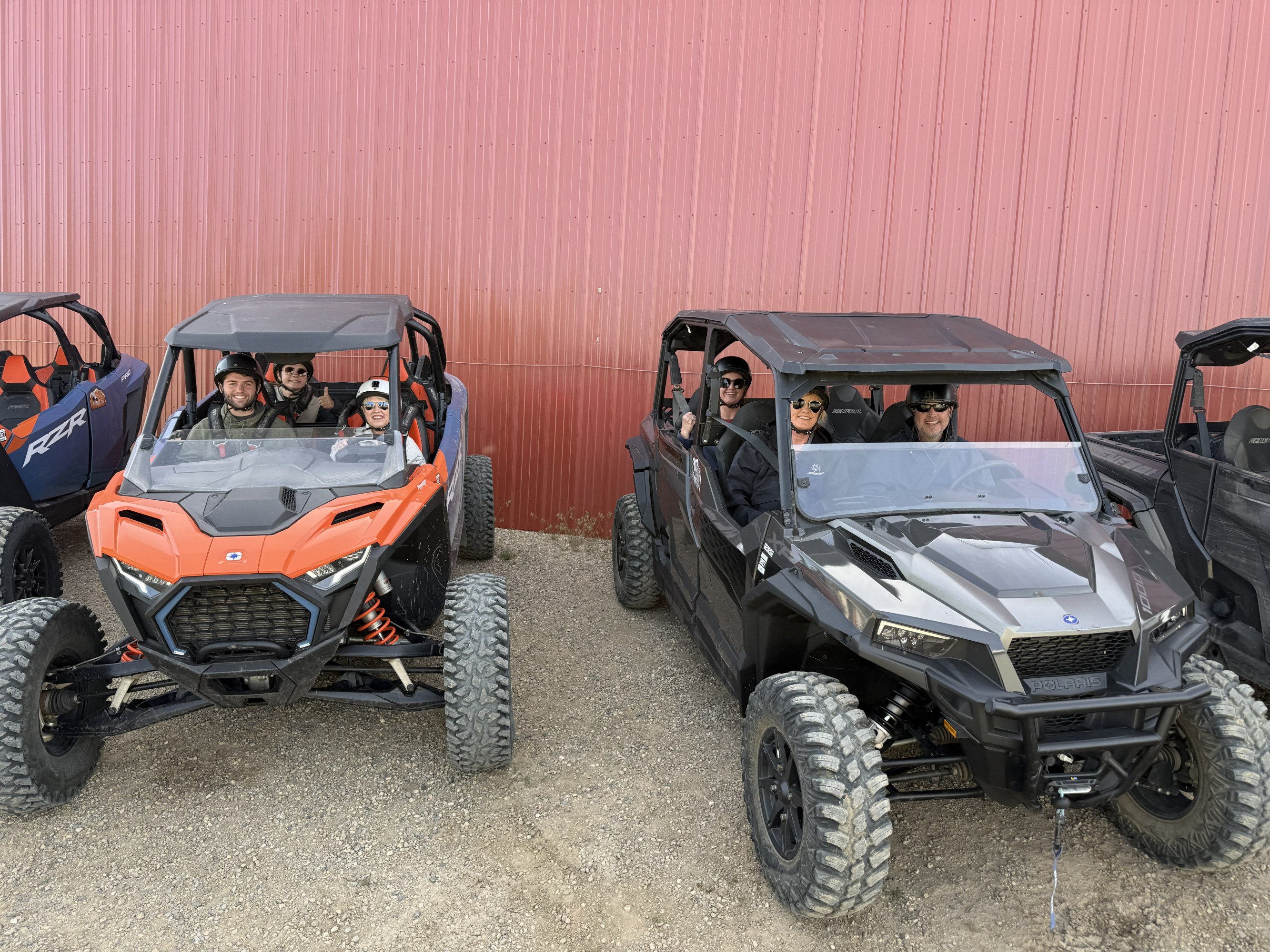 Two off-road vehicles with people inside parked in front of red wall.