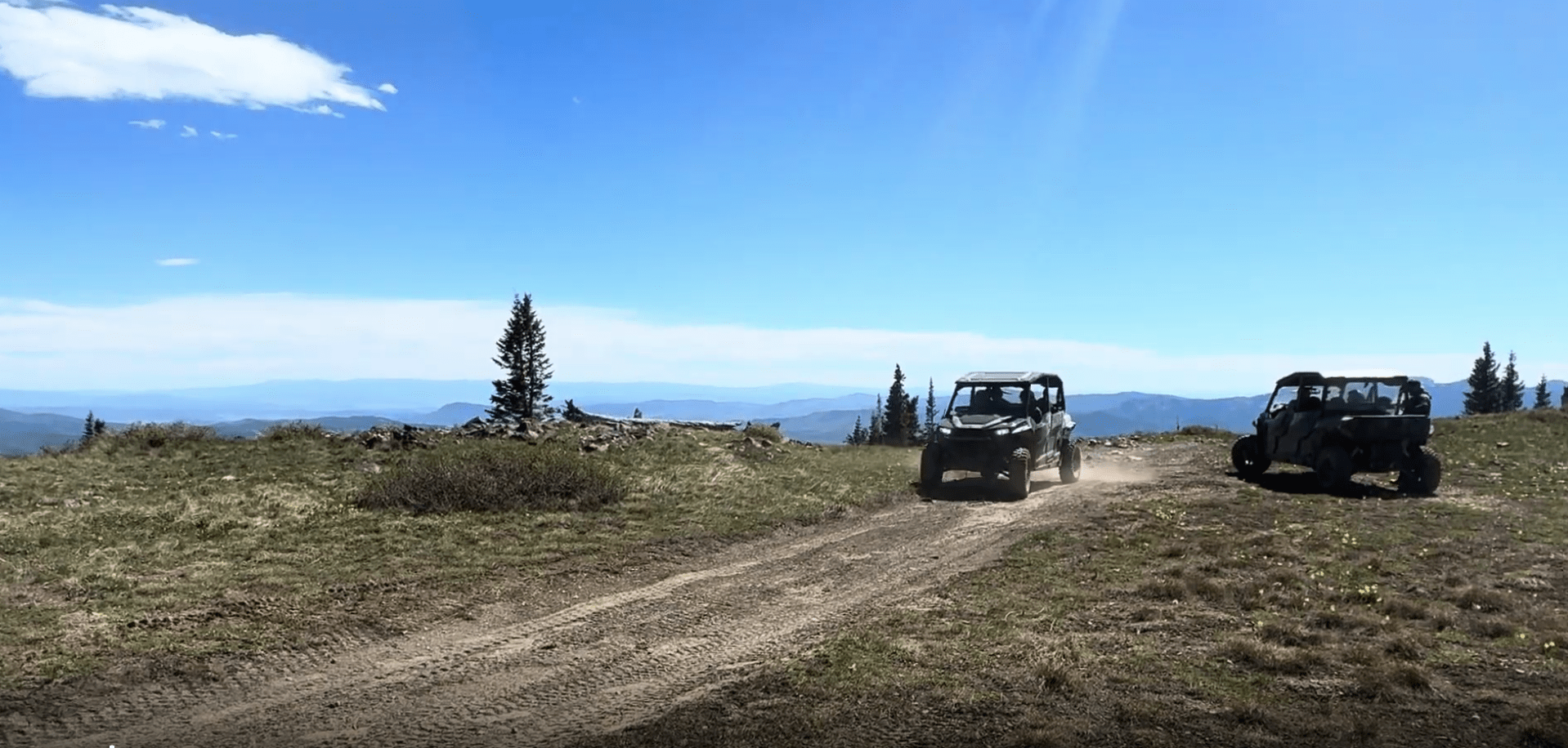 Two off-road vehicles on a dirt path in a mountain landscape under a clear blue sky.