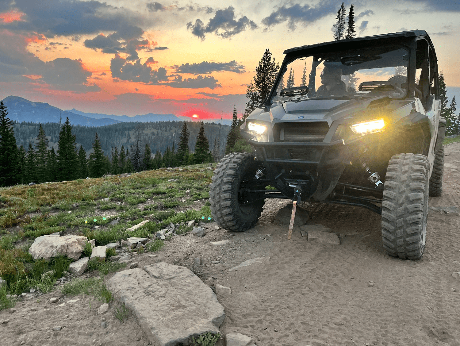 ATV on mountain trail at sunset with forest and mountains in background.